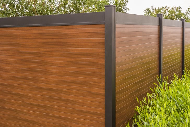 Close-up of an aluminum fence with a rich wood-like finish, featuring sleek, modern design elements and lush greenery in the background